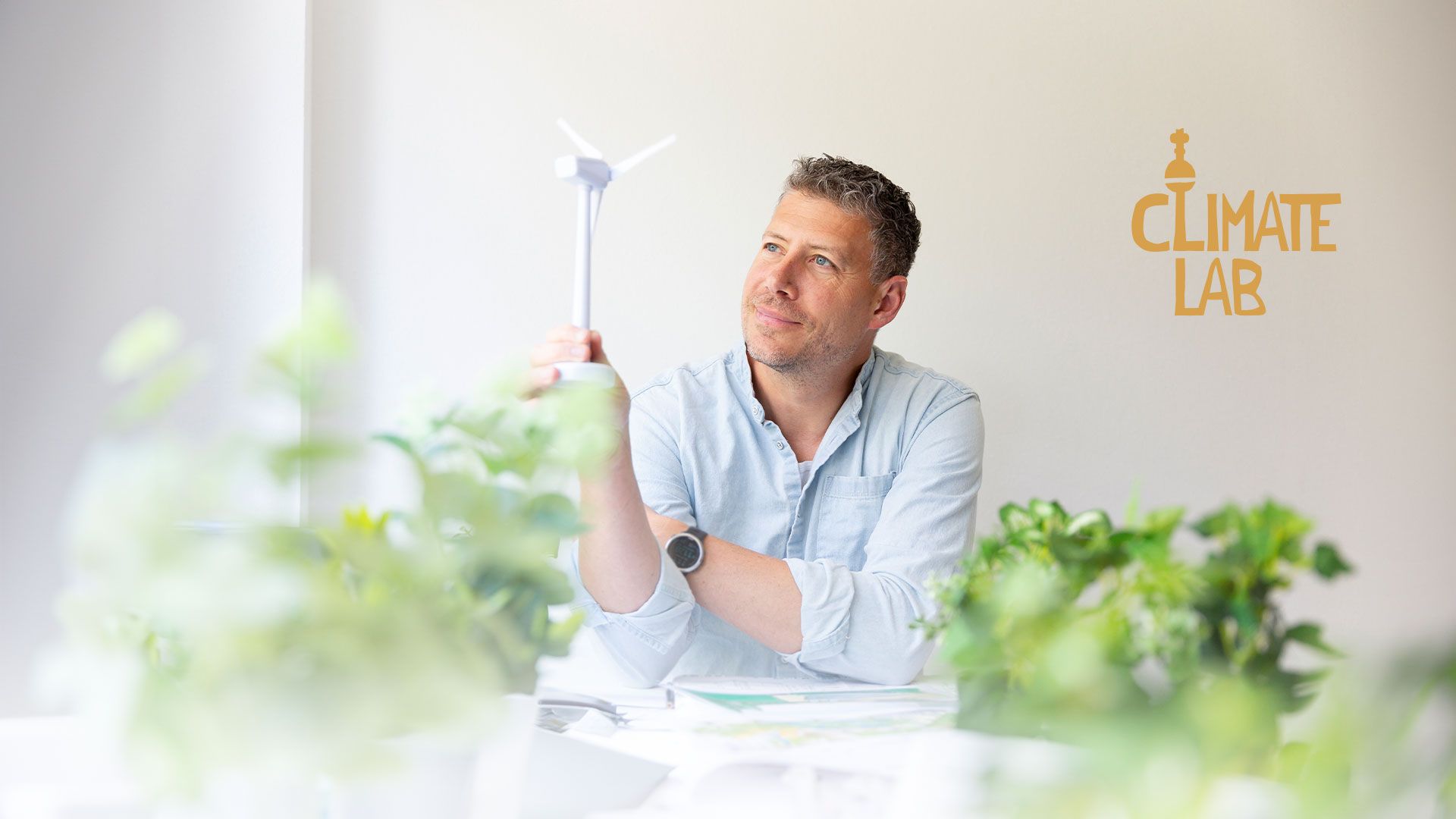 Mann mit hellblauem Hemd und Smartwatch blickt nachdenklich auf Windkraftanlage mit dem Logo 'Climate Lab' im Hintergrund