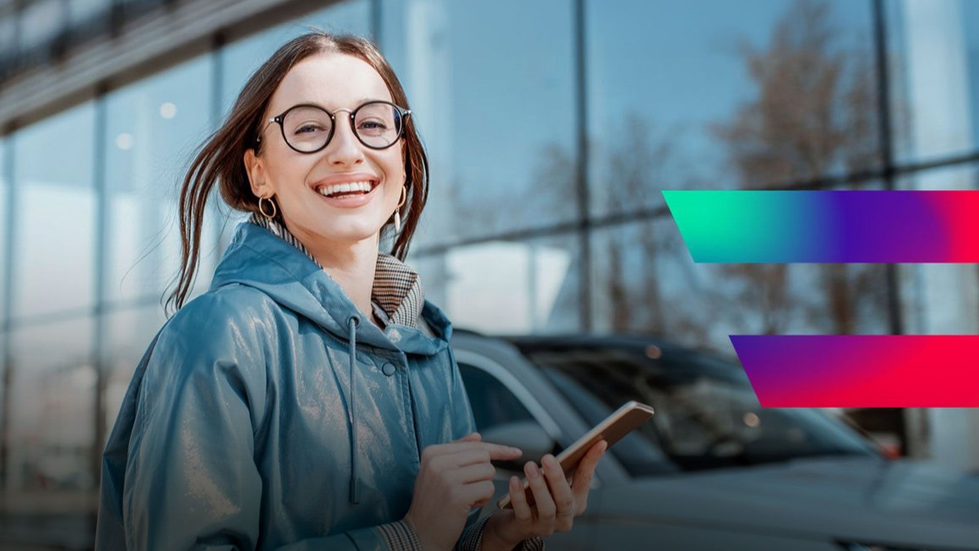 Junge Frau mit Brille und blauer Jacke lächelt und hält ein Smartphone vor einem modernen Gebäude