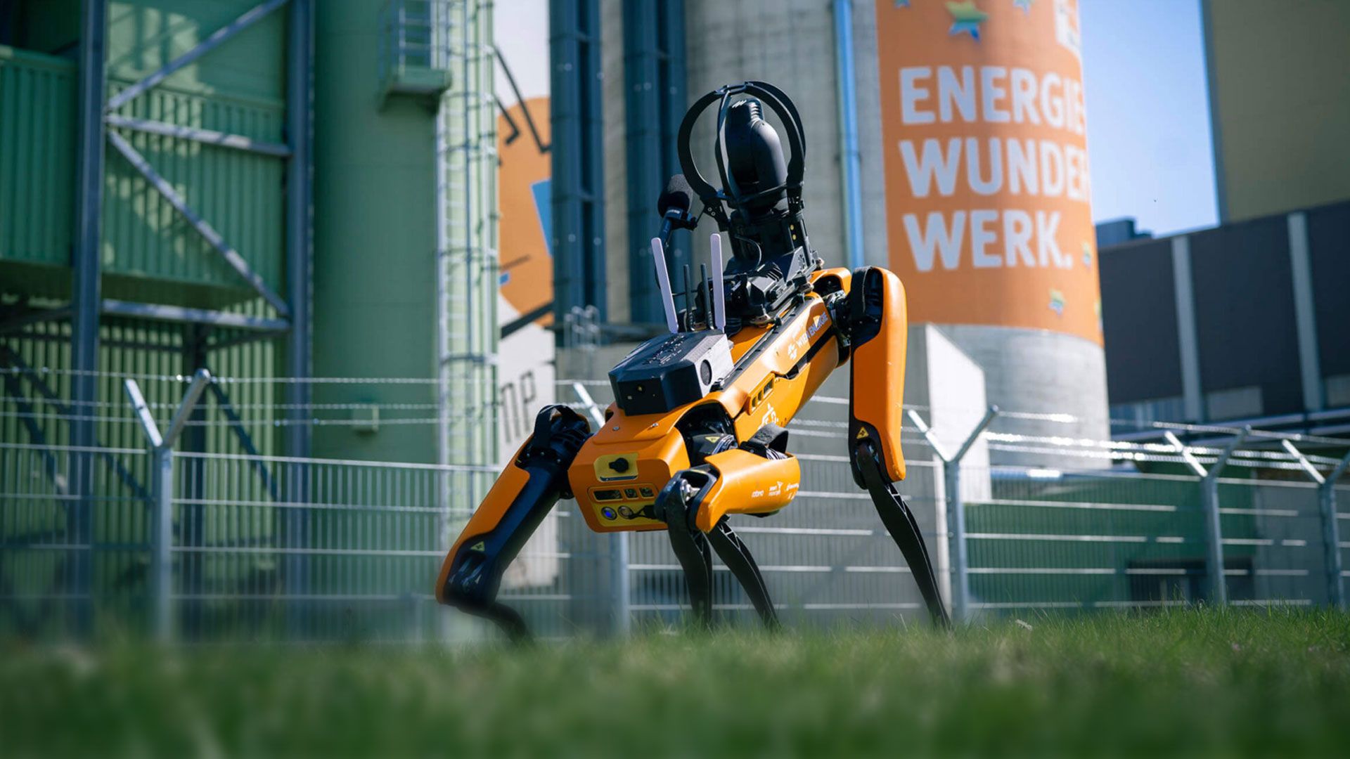 Oranger Roboterhund auf Gras vor Industrieanlage mit großem Zylinder, auf dem 'ENERGIE WUNDER WERK' steht