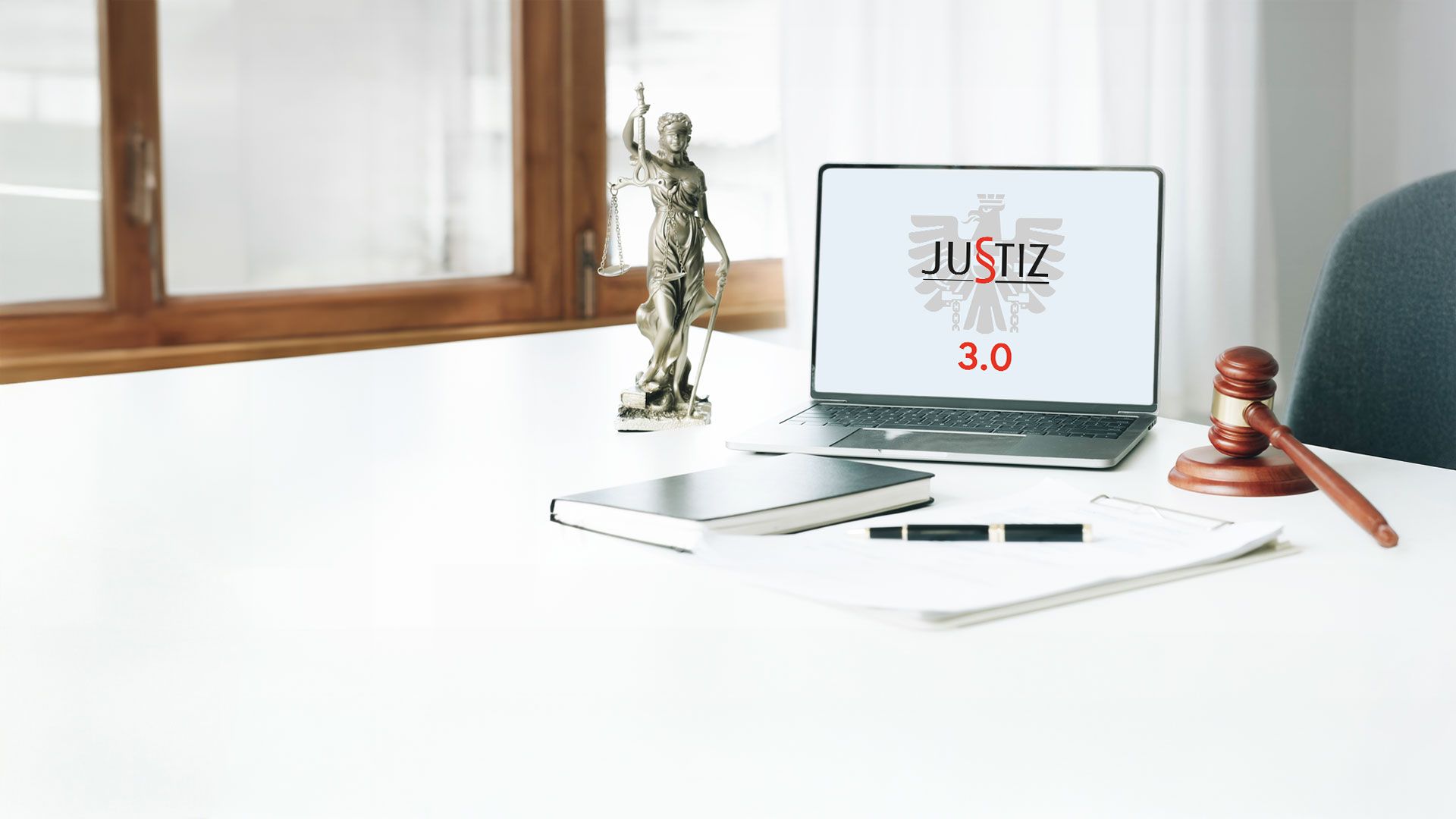 Laptop mit Justiz 3.0-Logo auf einem Schreibtisch, umgeben von Notizblock, Gerechtigkeitsfigur und Richterhammer.