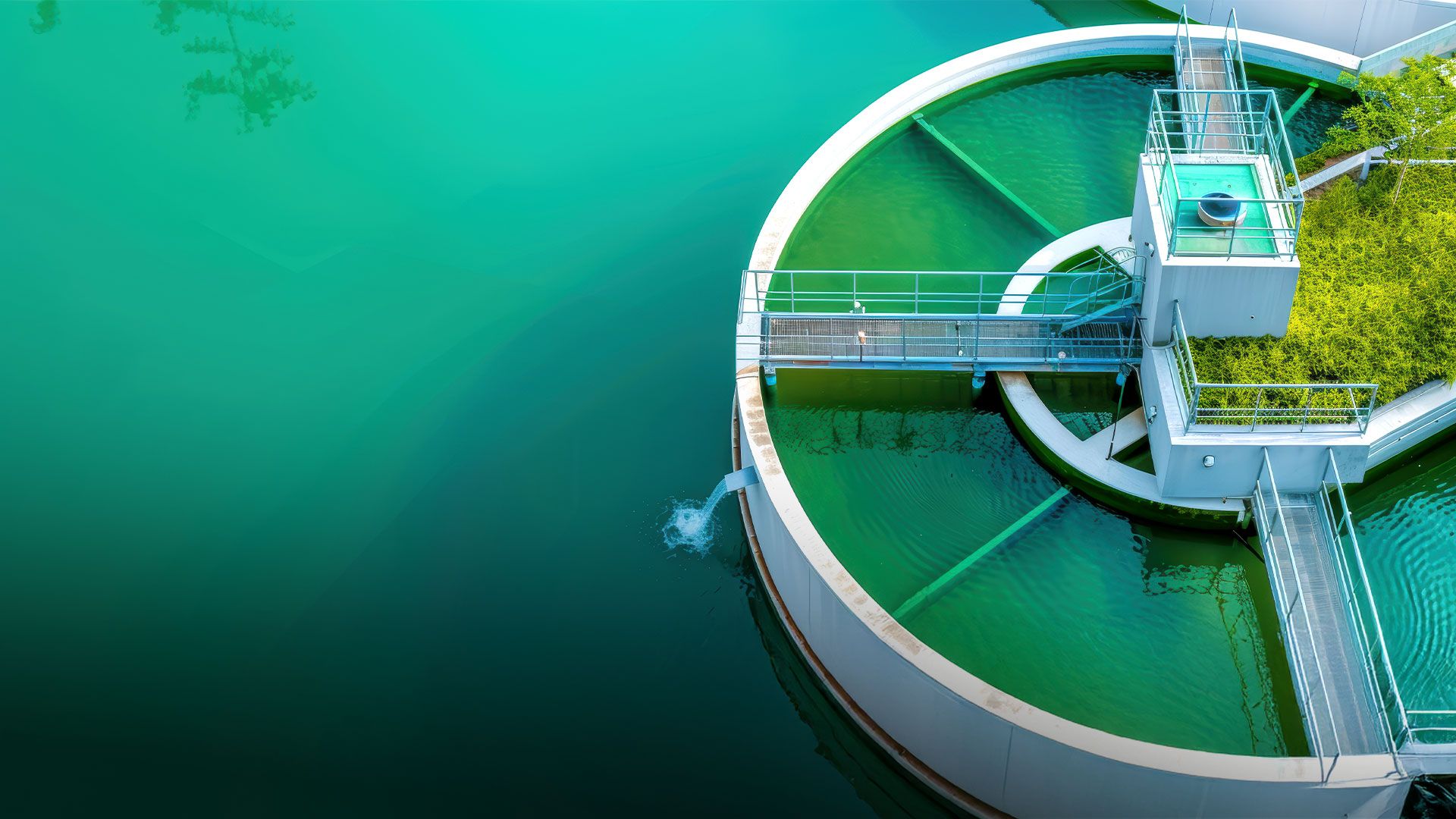 Luftaufnahme eines runden Klärbecken mit grünem Wasser und einer Brücke, die zur Mitte führt