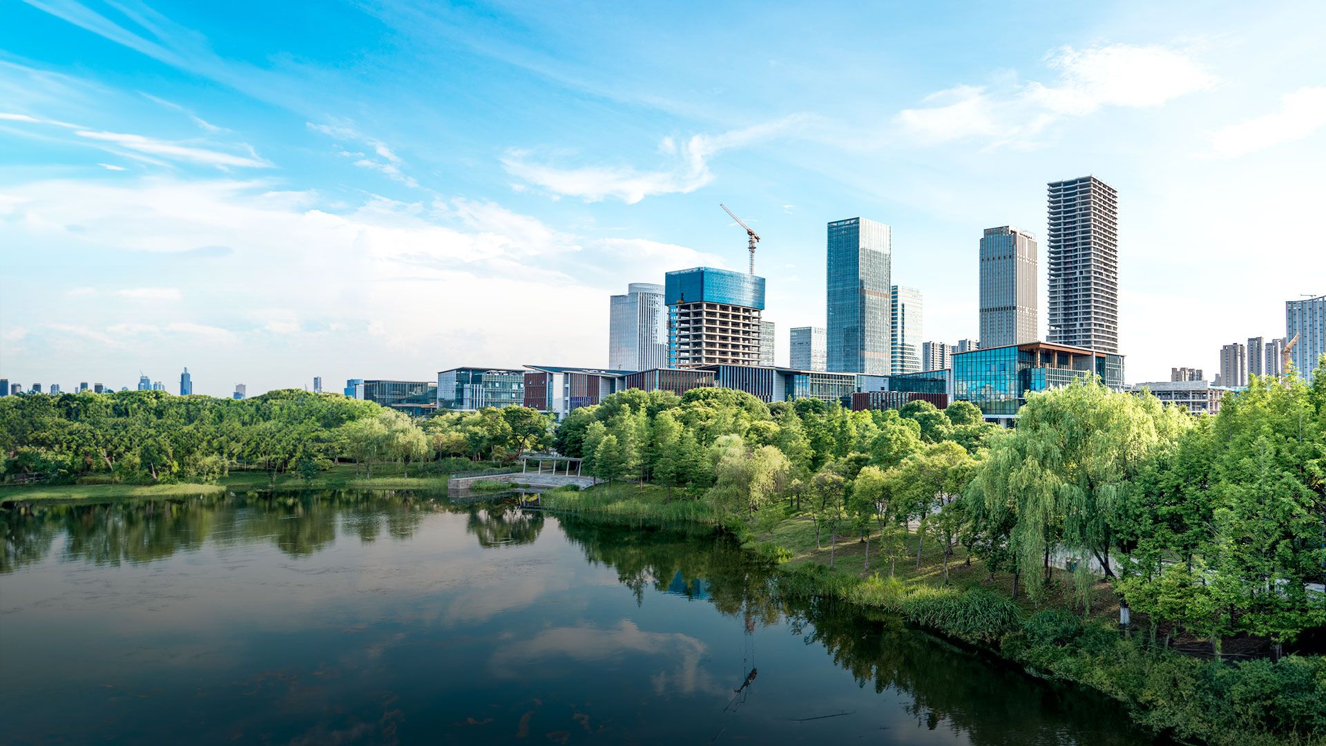 Moderne Skyline mit Hochhäusern und einem Kran im Bau, im Vordergrund ein grüner Park mit Bäumen und einem Teich, unter klarem, blauem Himmel.