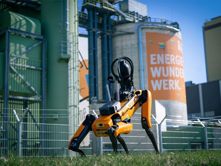 Oranger Roboterhund auf Gras vor Industrieanlage mit großem Zylinder, auf dem 'ENERGIE WUNDER WERK' steht