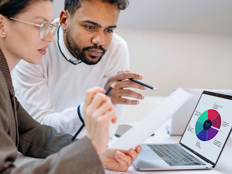 Zwei Personen analysieren gemeinsam ein Dokument vor einem Laptop mit einem farbigen Kreisdiagramm auf dem Bildschirm