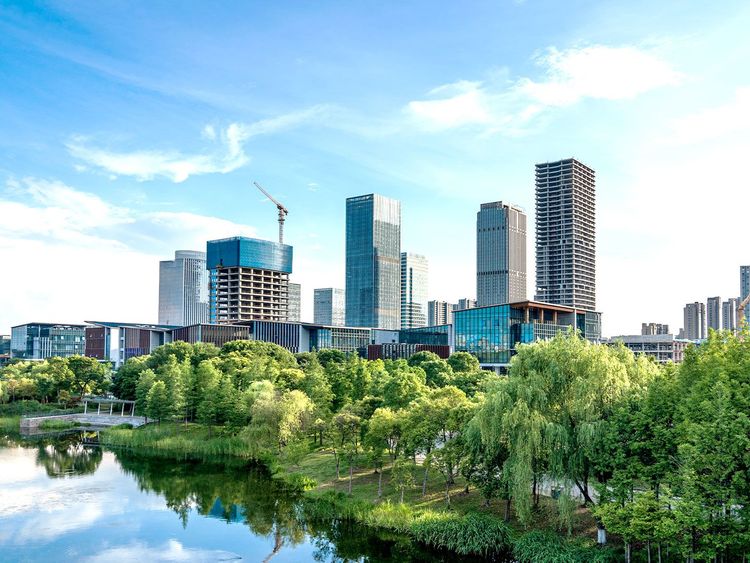 Moderne Skyline mit Hochhäusern und einem Kran im Bau, im Vordergrund ein grüner Park mit Bäumen und einem Teich, unter klarem, blauem Himmel.