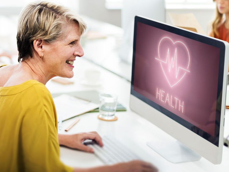 Ältere Frau lächelt am Schreibtisch vor Computerbildschirm mit Herz und Herzfrequenzsymbol und dem Wort 'HEALTH'