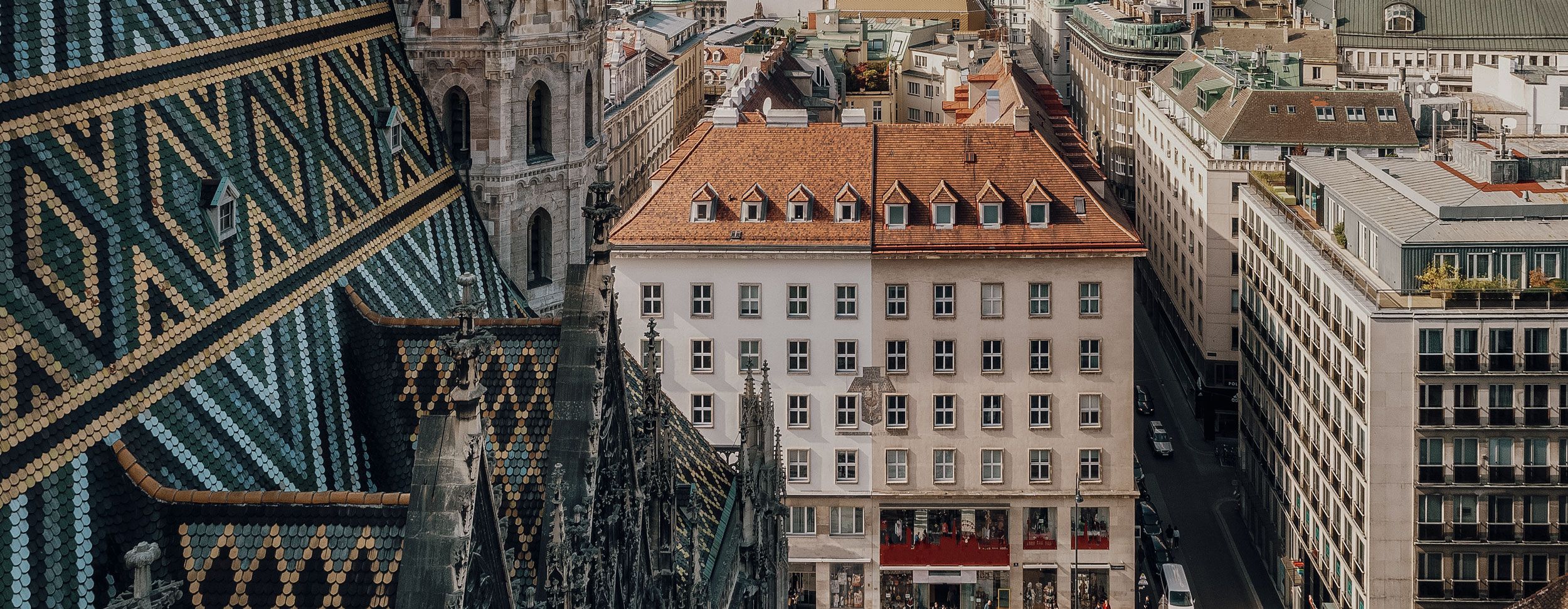 Dach mit bunten Ziegelmustern links, weißes Gebäude mit roten Fensterläden und rotem Dach in der Bildmitte, umgeben von weiteren Gebäuden und Straßen.